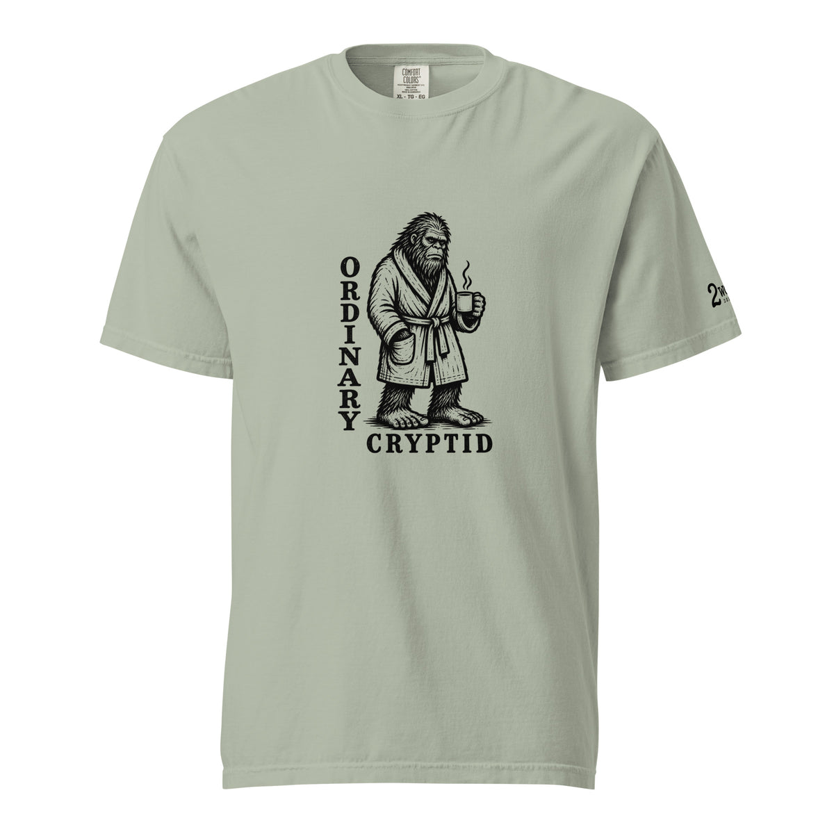 Black ink illustration of Bigfoot wearing a tied bathrobe and holding a steaming mug above the words “Ordinary Cryptid,” shown on a unisex t-shirt.