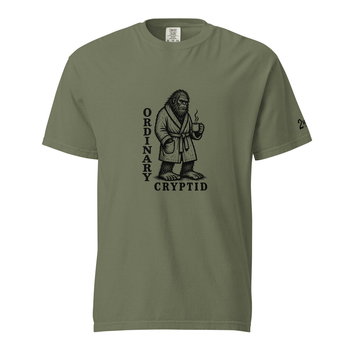 Black ink illustration of Bigfoot wearing a tied bathrobe and holding a steaming mug above the words “Ordinary Cryptid,” shown on a unisex t-shirt.