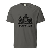 Black ink folk art illustration of a wide-eyed frog beneath an oversized pointed witch hat surrounded by cattails and marsh plants, with the words “BOG WITCH,” on a T-shirt.