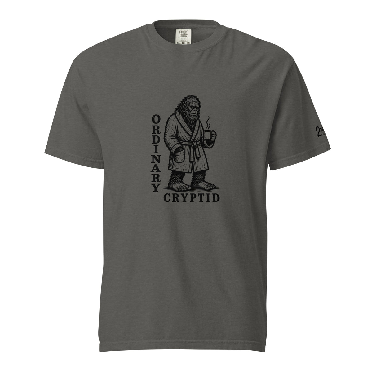Black ink illustration of Bigfoot wearing a tied bathrobe and holding a steaming mug above the words “Ordinary Cryptid,” shown on a unisex t-shirt.