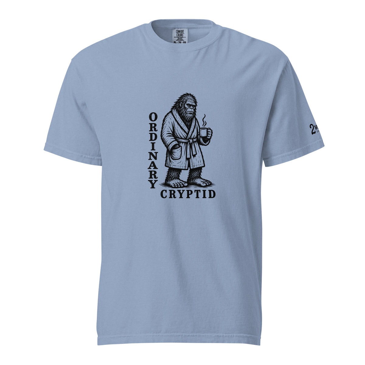 Black ink illustration of Bigfoot wearing a tied bathrobe and holding a steaming mug above the words “Ordinary Cryptid,” shown on a unisex t-shirt.