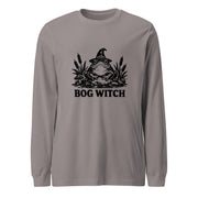 Black ink folk art illustration of a wide-eyed frog beneath an oversized pointed witch hat surrounded by cattails and marsh plants, with the words “BOG WITCH,” on a long sleeve tee.