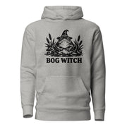 Black ink folk art illustration of a wide-eyed frog beneath an oversized pointed witch hat surrounded by cattails and marsh plants, with the words “BOG WITCH,” on a hoodie.