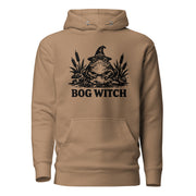 Black ink folk art illustration of a wide-eyed frog beneath an oversized pointed witch hat surrounded by cattails and marsh plants, with the words “BOG WITCH,” on a hoodie.