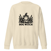 Black ink folk art illustration of a wide-eyed frog beneath an oversized pointed witch hat surrounded by cattails and marsh plants, with the words “BOG WITCH,” on a sweatshirt.