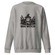 Black ink folk art illustration of a wide-eyed frog beneath an oversized pointed witch hat surrounded by cattails and marsh plants, with the words “BOG WITCH,” on a sweatshirt.