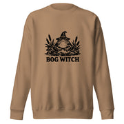 Black ink folk art illustration of a wide-eyed frog beneath an oversized pointed witch hat surrounded by cattails and marsh plants, with the words “BOG WITCH,” on a sweatshirt.