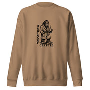 Black ink illustration of Bigfoot wearing a tied bathrobe and holding a steaming mug above the words “Ordinary Cryptid,” shown on a sweatshirt.